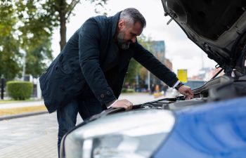 A car with open bonnet parked in a city street and a concerned man checking on fluids and parts.