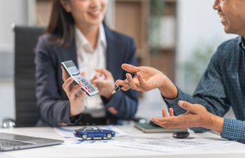 Two people sit at a desk discussing a car purchase. One holds a calculator, and the other gestures with a pen. A toy car and documents are on the table.