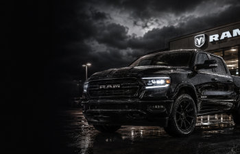 Black Ram pickup truck parked on wet pavement at night outside a dealership with the RAM logo illuminated under a dramatic cloudy sky.